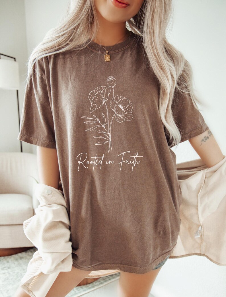 Puede incluir: Una camiseta marr&oacute;n de gran tama&ntilde;o con un contorno blanco de flores y el texto "Rooted in Faith".
