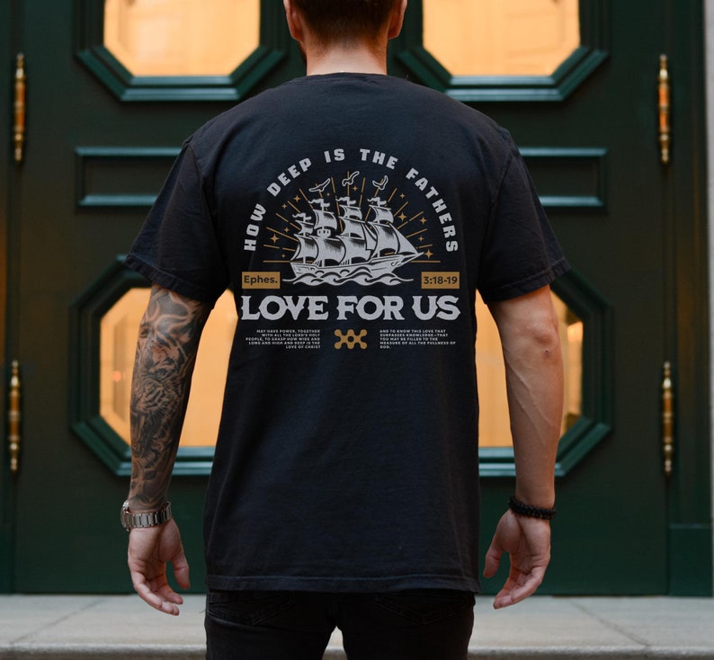 May include: Black t-shirt with a graphic design on the back. The design features a sailing ship, the text "HOW DEEP IS THE FATHERS LOVE FOR US", and the Bible verse Ephesians 3:18-19. The shirt is worn by a person.