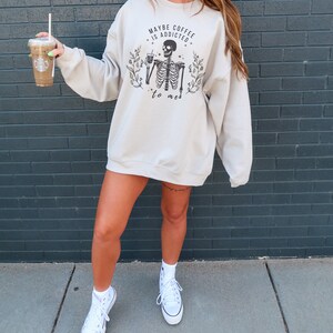 May include: A light beige sweatshirt with a skeleton graphic holding a cup of coffee. The text on the sweatshirt reads "Maybe coffee is addicted to me".