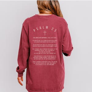 Może przedstawiać: Bordowy t-shirt z długim rękawem z białym nadrukiem Psalmu 23, wersetu chrześcijańskiego. Tekst zawiera słowa "Pan jest moim pasterzem, niczego mi nie zabraknie".