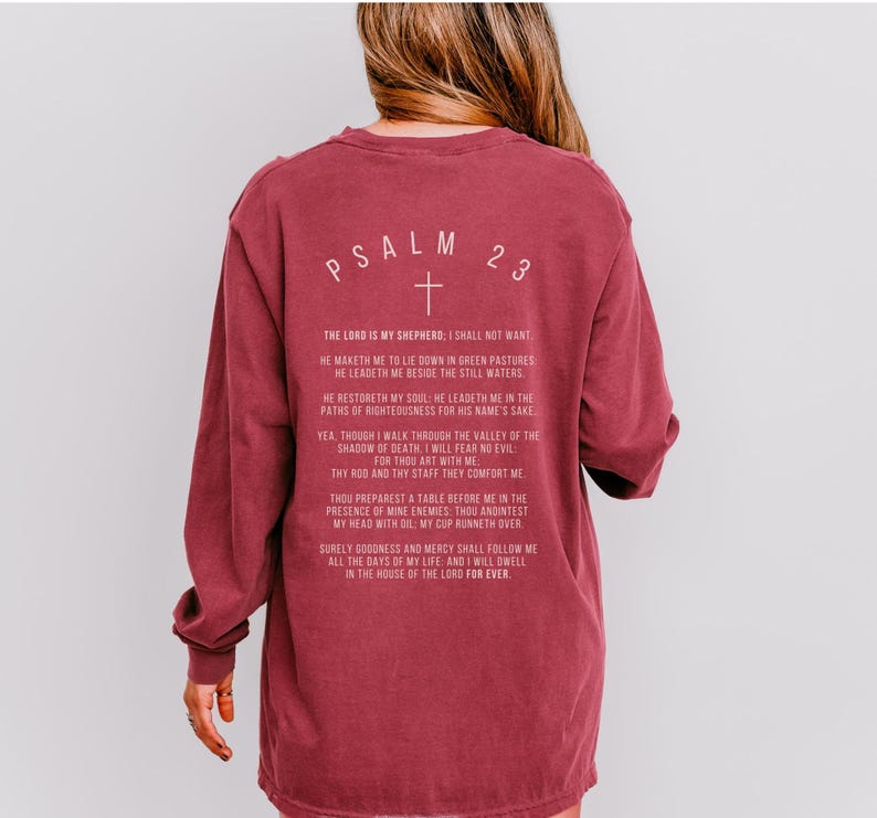 Może przedstawiać: Bordowy t-shirt z długim rękawem z białym nadrukiem Psalmu 23, wersetu chrześcijańskiego. Tekst zawiera słowa "Pan jest moim pasterzem, niczego mi nie zabraknie".