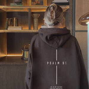 May include: A brown hoodie with the text "PSALM 91" and a vertical line design. Below the text is the quote "HE IS MY REFUGE AND MY FORTRESS, MY GOD, IN WHOM I TRUST" in white. The hoodie is worn by a person.