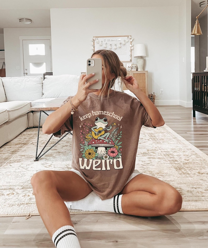 May include: Brown t-shirt with the text "keep homeschool weird" and a graphic of a frog playing a guitar on a mushroom. The shirt is worn with white shorts and striped socks. The person is holding a phone.