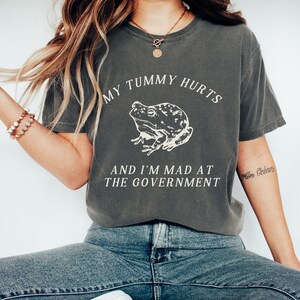 May include: A dark gray t-shirt with a white graphic of a frog and the text "My tummy hurts and I'm mad at the government".
