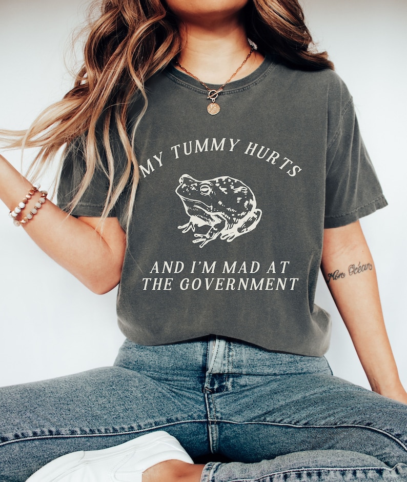 May include: A dark gray t-shirt with a white graphic of a frog and the text "My tummy hurts and I'm mad at the government".
