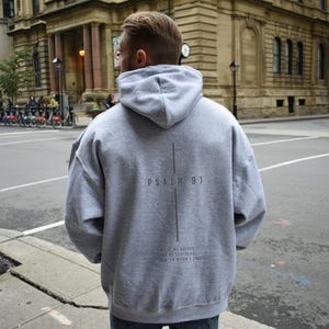 May include: A gray hoodie with the text "Psalm 91" and a vertical line design on the back. Below the text is the phrase "He is my refuge and my fortress, in whom I trust." The hoodie has a hood and a relaxed fit.