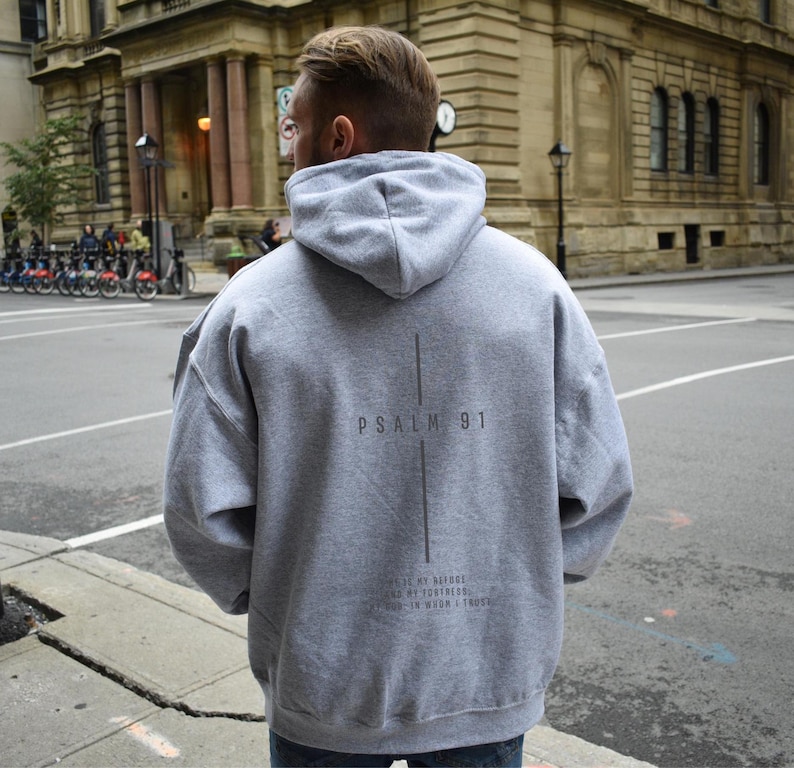 May include: A gray hoodie with the text "Psalm 91" and a vertical line design on the back. Below the text is the phrase "He is my refuge and my fortress, in whom I trust." The hoodie has a hood and a relaxed fit.