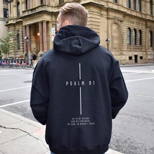 May include: Black hooded sweatshirt with the text "PSALM 91" and a vertical cross design on the back. Below the text is the phrase "HE IS MY REFUGE AND MY FORTRESS, MY GOD, IN WHOM I TRUST" in white.