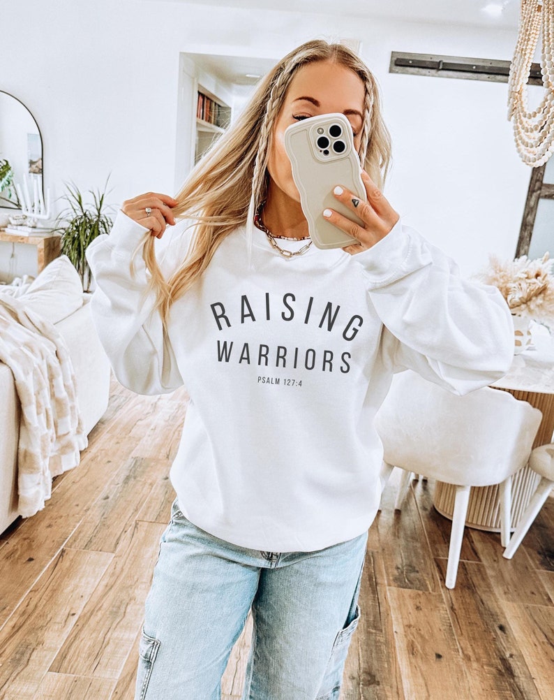 May include: White sweatshirt with the text "RAISING WARRIORS" and "PSALM 127:4" in black. The sweatshirt is worn with light blue jeans. The image is taken indoors.