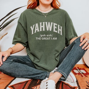 Op de afbeelding: Een groen T-shirt met het woord "YAHWEH" in witte tekst met een vintage lettertype. De tekst "yah-weh" staat in kleinere letters onder het woord "YAHWEH". De tekst "THE GREAT I AM" staat onder het woord "yah-weh".