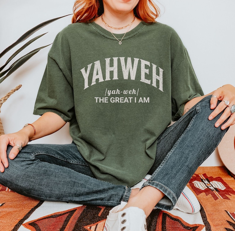 Op de afbeelding: Een groen T-shirt met het woord "YAHWEH" in witte tekst met een vintage lettertype. De tekst "yah-weh" staat in kleinere letters onder het woord "YAHWEH". De tekst "THE GREAT I AM" staat onder het woord "yah-weh".