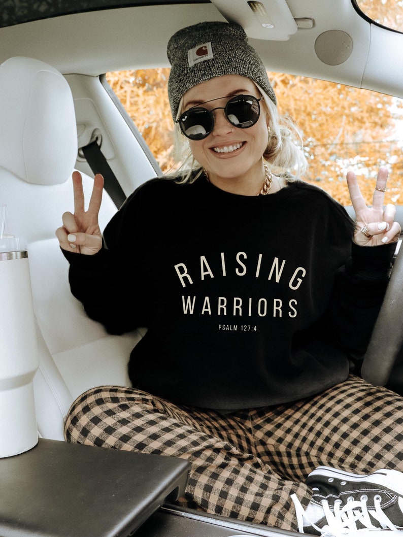 May include: A black sweatshirt with the text "RAISING WARRIORS" and "PSALM 127:4" in white. The person is wearing a grey beanie, black sunglasses, and a gold chain necklace. They are sitting in a car and wearing plaid trousers.