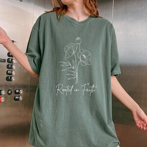 Puede incluir: Una camiseta verde de gran tama&ntilde;o con un contorno blanco de una flor y el texto "Rooted in Faith".
