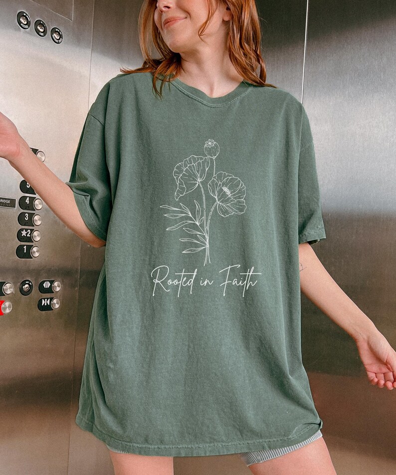 Puede incluir: Una camiseta verde de gran tama&ntilde;o con un contorno blanco de una flor y el texto "Rooted in Faith".