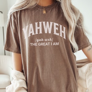 Op de afbeelding: Een bruin T-shirt met het woord "YAHWEH" in witte letters met een roze omtrek. De tekst "yah-weh" staat in kleinere letters onder het woord "YAHWEH". De tekst "THE GREAT I AM" staat in kleinere letters onder het woord "yah-weh".