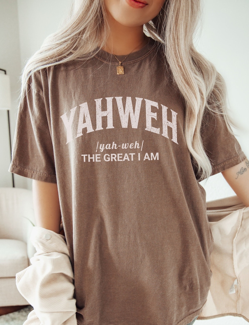 Op de afbeelding: Een bruin T-shirt met het woord "YAHWEH" in witte letters met een roze omtrek. De tekst "yah-weh" staat in kleinere letters onder het woord "YAHWEH". De tekst "THE GREAT I AM" staat in kleinere letters onder het woord "yah-weh".