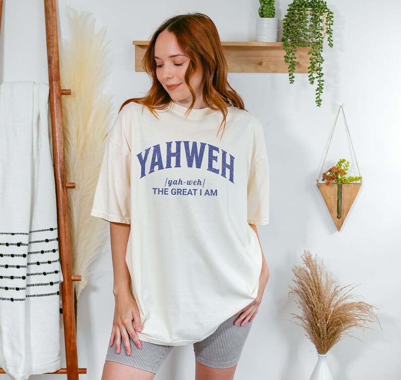 Op de afbeelding: Een cr&egrave;mekleurig T-shirt met een blauwe afbeelding waarop "YAHWEH /yah-weh/ THE GREAT I AM" staat.