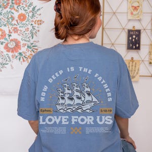 May include: A light blue t-shirt with the text "HOW DEEP IS THE FATHERS LOVE FOR US" and a graphic of a ship. The shirt also includes the text "Ephes. 3:18-19" and additional text below the ship graphic.