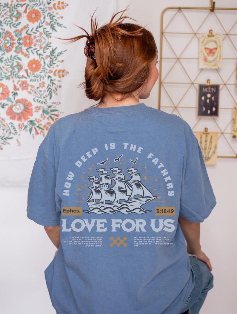 May include: A light blue t-shirt with the text "HOW DEEP IS THE FATHERS LOVE FOR US" and a graphic of a ship. The shirt also includes the text "Ephes. 3:18-19" and additional text below the ship graphic.