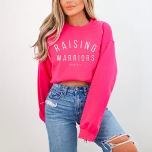 May include: A vibrant pink sweatshirt featuring the white text "RAISING WARRIORS" in an arc, with "PSALM 127:4" underneath. The sweatshirt is worn with blue denim jeans.