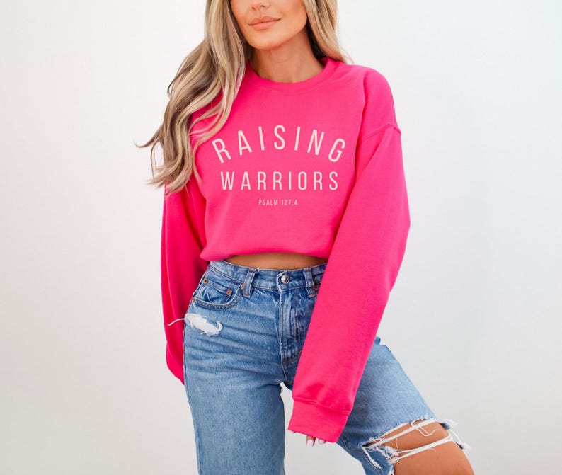 May include: A vibrant pink sweatshirt featuring the white text "RAISING WARRIORS" in an arc, with "PSALM 127:4" underneath. The sweatshirt is worn with blue denim jeans.