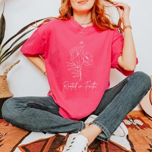 Puede incluir: Una camiseta rosa con un contorno blanco de flores y el texto "Rooted in Faith".