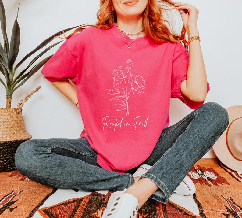 Puede incluir: Una camiseta rosa con un contorno blanco de flores y el texto "Rooted in Faith".