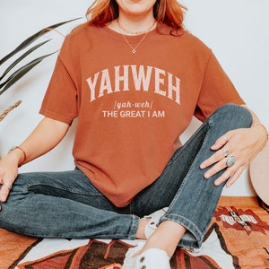 Op de afbeelding: Een roestkleurig T-shirt met het woord "YAHWEH" in witte tekst met een vintage verweerde look. De tekst "yah-weh" en "THE GREAT I AM" staan in een kleiner lettertype onder het woord "YAHWEH".