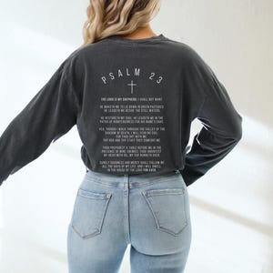 Może przedstawiać: Czarny, długorękawny t-shirt z białym nadrukiem Psalmu 23, wersetu biblijnego. T-shirt jest noszony przez osobę w niebieskich dżinsach.