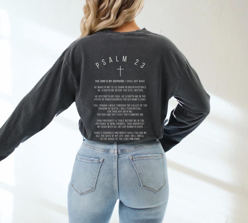 Może przedstawiać: Czarny, długorękawny t-shirt z białym nadrukiem Psalmu 23, wersetu biblijnego. T-shirt jest noszony przez osobę w niebieskich dżinsach.