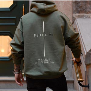 May include: Olive green hoodie with the text "PSALM 91" and a vertical line design on the back. Below the text is the quote "HE IS MY REFUGE AND MY FORTRESS. MY GOD, IN WHOM I TRUST."
