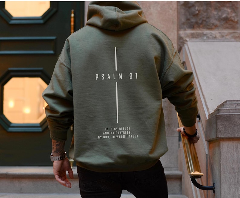 May include: Olive green hoodie with the text "PSALM 91" and a vertical line design on the back. Below the text is the quote "HE IS MY REFUGE AND MY FORTRESS. MY GOD, IN WHOM I TRUST."