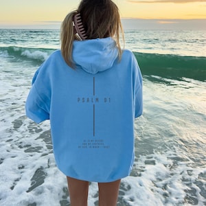 May include: A light blue hoodie with the text "PSALM 91" and a verse printed on the back. The hoodie is worn by a person standing in the ocean. The sky is a mix of yellow and orange.