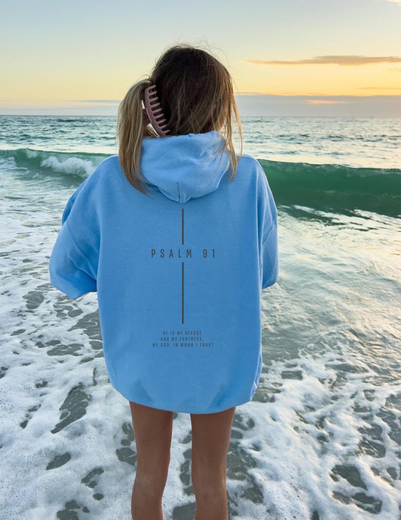 May include: A light blue hoodie with the text "PSALM 91" and a verse printed on the back. The hoodie is worn by a person standing in the ocean. The sky is a mix of yellow and orange.