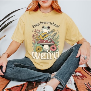 May include: Yellow t-shirt with a whimsical graphic of a frog playing a guitar on a mushroom, surrounded by flowers. The text reads "keep homeschool weird". The shirt is worn by a person.