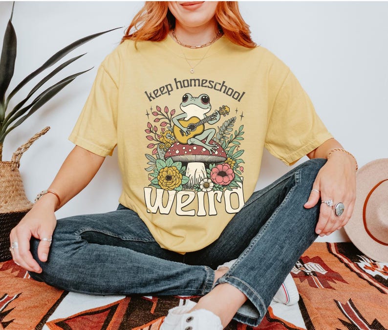 May include: Yellow t-shirt with a whimsical graphic of a frog playing a guitar on a mushroom, surrounded by flowers. The text reads "keep homeschool weird". The shirt is worn by a person.