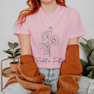 Puede incluir: Una camiseta rosa con un dibujo de flores en l&iacute;nea negra y el texto "Rooted in Faith".