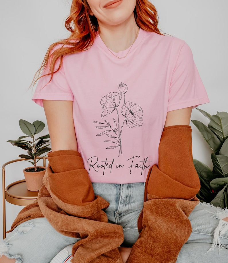 Puede incluir: Una camiseta rosa con un dibujo de flores en l&iacute;nea negra y el texto "Rooted in Faith".