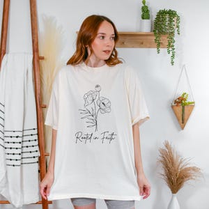 Puede incluir: Una camiseta blanca de gran tama&ntilde;o con un dibujo de l&iacute;nea negra de una flor y el texto "Rooted in Faith".