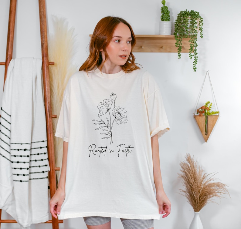 Puede incluir: Una camiseta blanca de gran tama&ntilde;o con un dibujo de l&iacute;nea negra de una flor y el texto "Rooted in Faith".