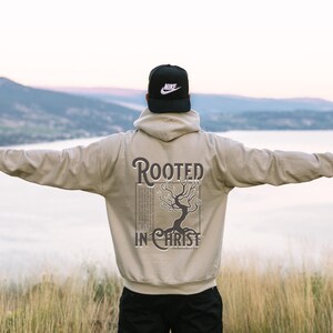 May include: A person wearing a beige hoodie with the text "Rooted in Christ" and a tree graphic on the back. The person is standing with their arms outstretched, facing away from the camera. The background is a blurry image of a lake and mountains.