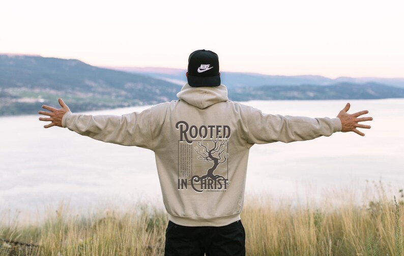 May include: A person wearing a beige hoodie with the text "Rooted in Christ" and a tree graphic on the back. The person is standing with their arms outstretched, facing away from the camera. The background is a blurry image of a lake and mountains.