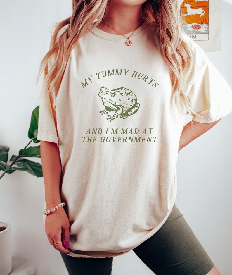 May include: A cream-colored t-shirt with a green frog graphic and the text "My tummy hurts and I'm mad at the government".