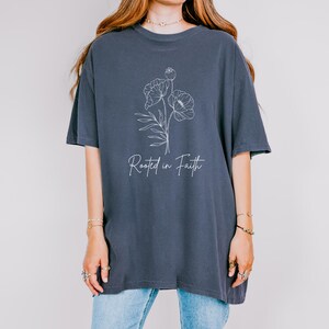 Puede incluir: Una camiseta de gran tama&ntilde;o de color azul oscuro con un contorno blanco de flores y el texto "Rooted in Faith".