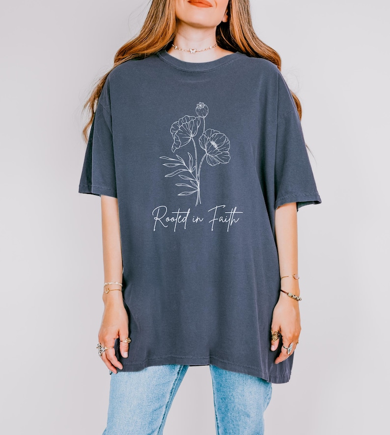 Puede incluir: Una camiseta de gran tama&ntilde;o de color azul oscuro con un contorno blanco de flores y el texto "Rooted in Faith".