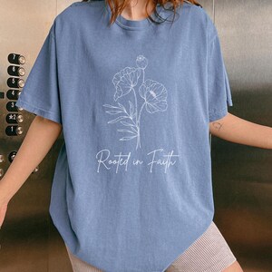 Puede incluir: Una camiseta azul claro con un contorno blanco de flores y el texto "Rooted in Faith".