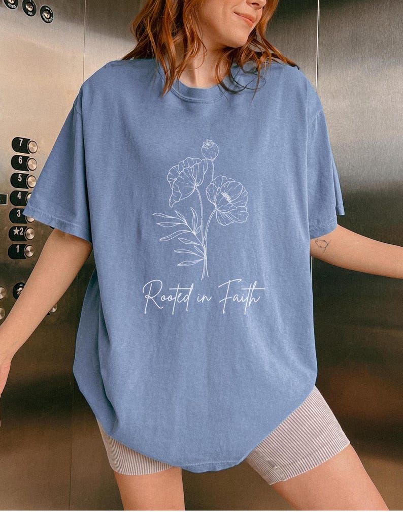 Puede incluir: Una camiseta azul claro con un contorno blanco de flores y el texto "Rooted in Faith".