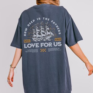 May include: Dark gray t-shirt with a graphic on the back. The graphic features a sailing ship with the text "HOW DEEP IS THE FATHERS LOVE FOR US" and the scripture reference "Ephes. 3:18-19". The design is in white and gold.