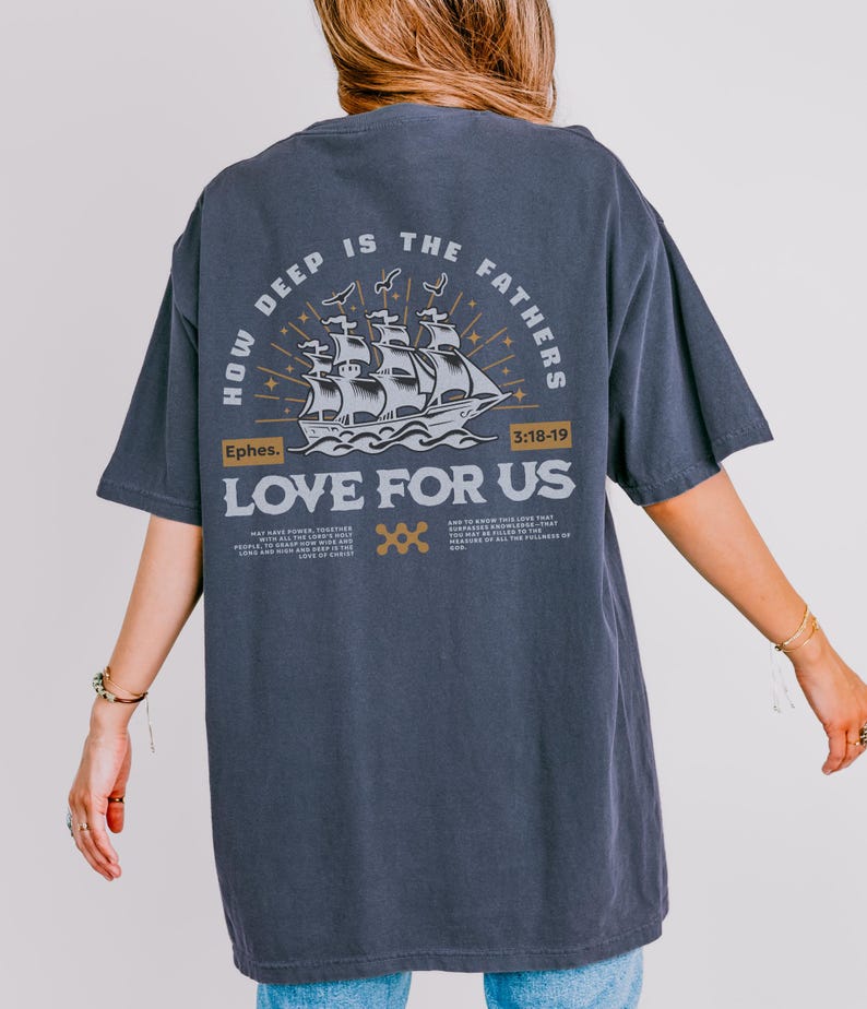 May include: Dark gray t-shirt with a graphic on the back. The graphic features a sailing ship with the text "HOW DEEP IS THE FATHERS LOVE FOR US" and the scripture reference "Ephes. 3:18-19". The design is in white and gold.