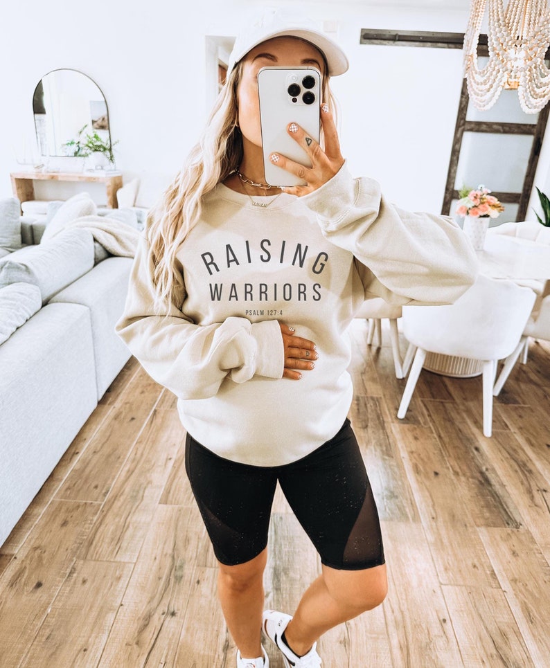 May include: A beige sweatshirt with the text "RAISING WARRIORS" in black, with "PSALM 127:4" below. The sweatshirt is worn with black cycling shorts and a white baseball cap. The outfit is completed with white trainers.
